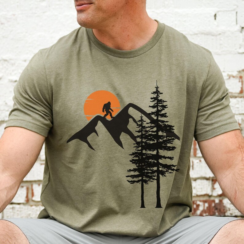 May include: A green t-shirt with a graphic of a Bigfoot walking on a mountain range with a sunset behind him. Two pine trees are in the foreground.