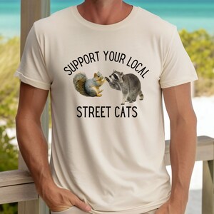 May include: A beige t-shirt with a black graphic of a raccoon and a squirrel shaking hands. The text "Support Your Local Street Cats" is printed below the graphic.