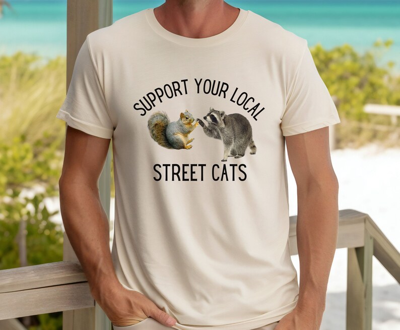 May include: A beige t-shirt with a black graphic of a raccoon and a squirrel shaking hands. The text "Support Your Local Street Cats" is printed below the graphic.