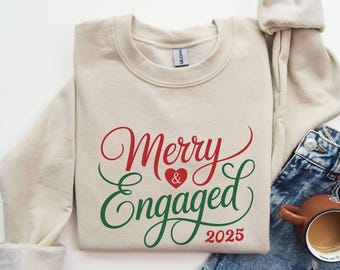 May include: A sand-colored sweatshirt with the words "Merry & Engaged 2025" in red and green script. The design includes a heart symbol. The sweatshirt is folded and placed on top of blue jeans.