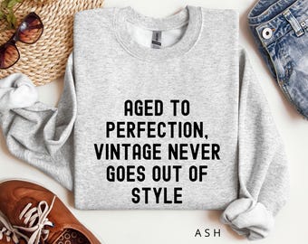 May include: Gray sweatshirt with the text "Aged to Perfection, Vintage Never Goes Out of Style" in black lettering. The text is centered on the sweatshirt. The brand name "ASH" is printed in black at the bottom of the sweatshirt.