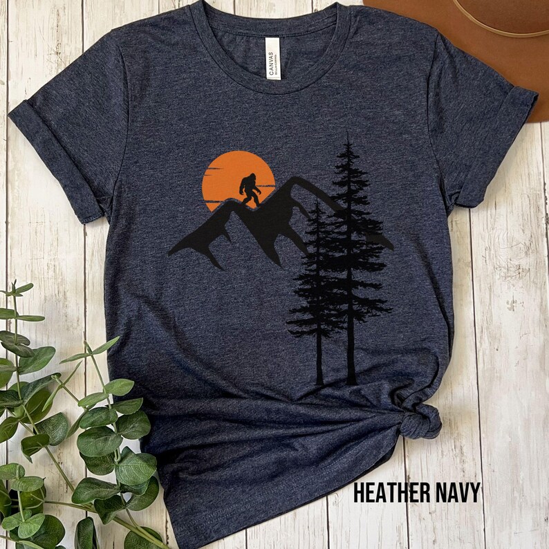 May include: A heather navy blue t-shirt with a graphic of a Bigfoot walking on a mountain range with a sunset behind him. The graphic also includes two pine trees.