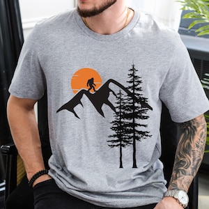 May include: Gray t-shirt with a black silhouette of a Bigfoot walking on a mountain range with a setting sun and two pine trees.
