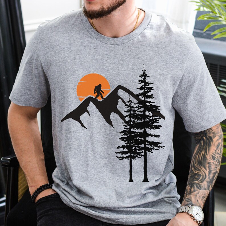May include: Gray t-shirt with a black silhouette of a Bigfoot walking on a mountain range with a setting sun and two pine trees.