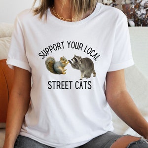 May include: White t-shirt with the text "Support Your Local Street Cats" printed on it. The text is in black and there is a graphic of a raccoon and a squirrel shaking paws.