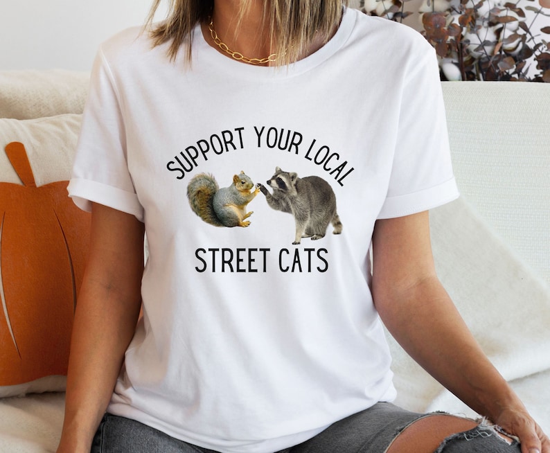 May include: White t-shirt with the text "Support Your Local Street Cats" printed on it. The text is in black and there is a graphic of a raccoon and a squirrel shaking paws.