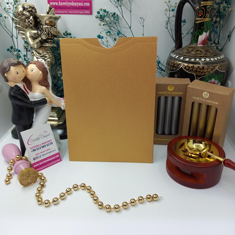 May include: A gold envelope with a flap on the top, a pink wax seal with a tree design, a string of gold beads, and two boxes of wax sticks. The boxes are labeled "Davetiye Dunyasi" which translates to "Invitation World".