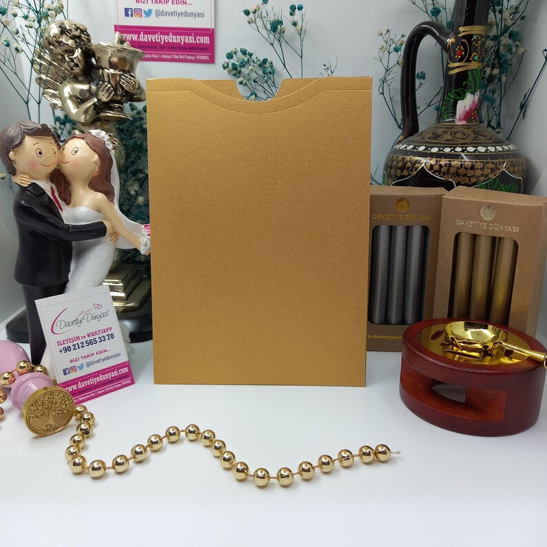 May include: Gold envelope with a flap on the top. The envelope is sitting on a table with a gold chain, a wax seal, and a wax seal kit with gold and silver wax sticks.