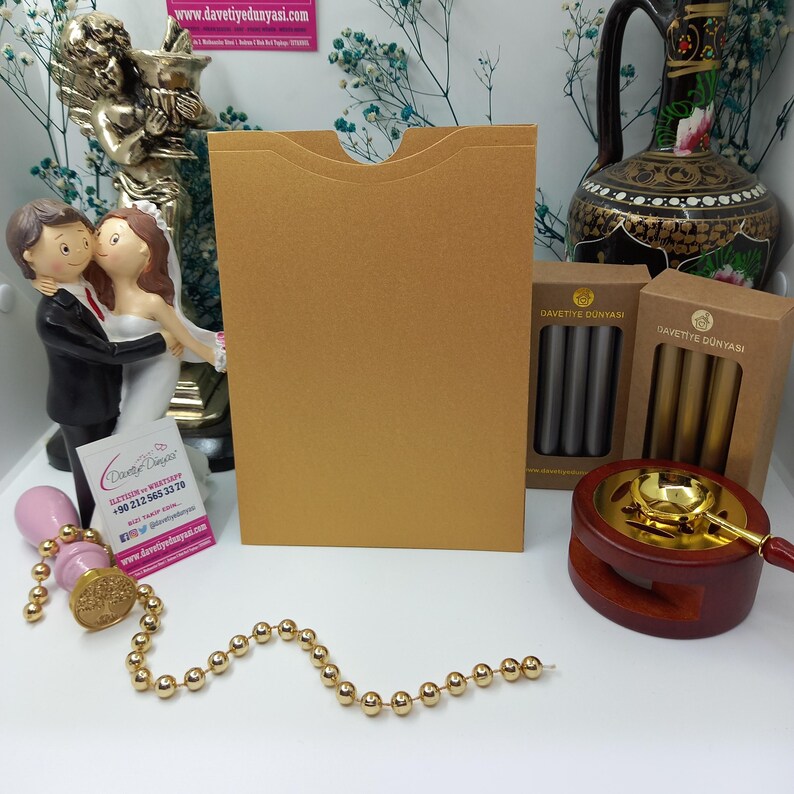 May include: A gold envelope with a pocket for a card, a wax seal kit with a wooden base, a gold chain, and two boxes of wax sticks labeled "Davetiye Dunyasi" in Turkish.