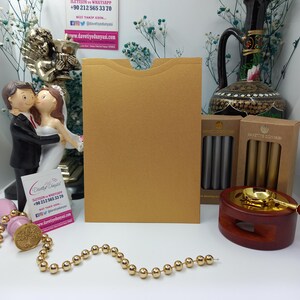 May include: A gold envelope with a flap on the top. The envelope is sitting on a white surface. There are two boxes of wax seals, one with gold seals and one with silver seals. There is also a wax seal stamp and a chain of gold beads.
