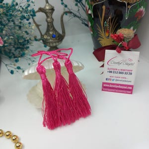 May include: Four pink tassel embellishments with a white string attached to each. The tassels are arranged in a row on a white surface. The text on the card reads "Davetiye Dunyasi", "Iletisim ve Whatsapp", "+90 212 565 33 70", "Bizi Takip Edin...", "@davetiyedunyasi", "www.davetiyedunyasi.com".