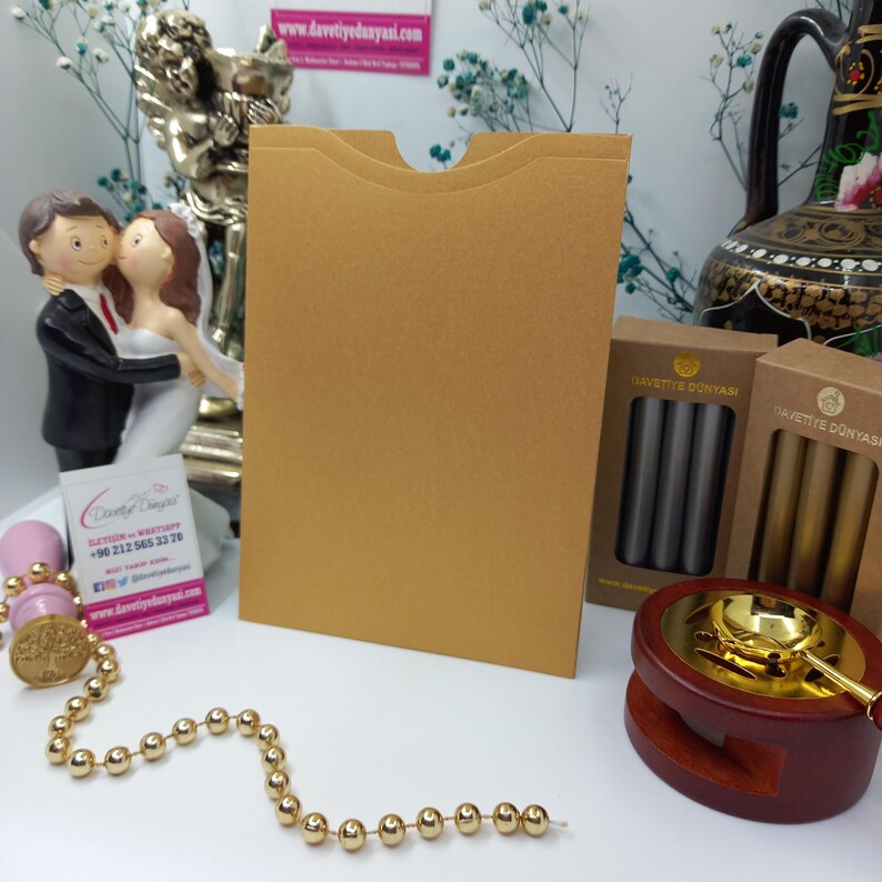 May include: A gold envelope with a flap on the top. The envelope is sitting on a table with a wax seal kit, a string of gold beads, and a business card.