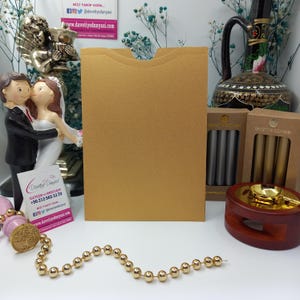 May include: A gold envelope with a flap on the top. The envelope is sitting on a table with a small wooden stand, a gold and silver wax seal kit, and a pair of wedding figurines.