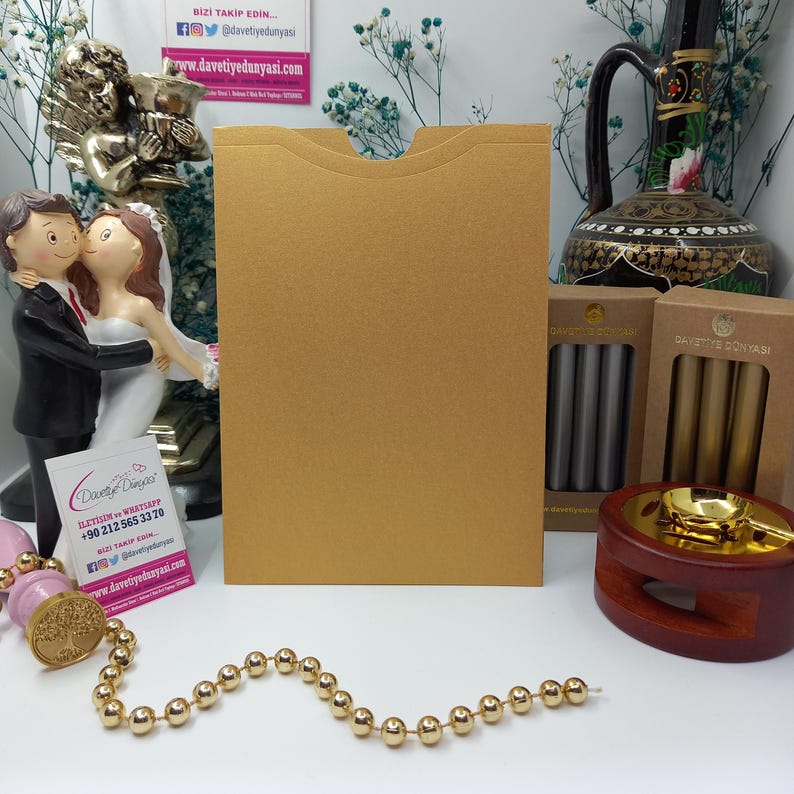May include: A gold envelope with a flap on the top. The envelope is sitting on a table with a small wooden stand, a gold and silver wax seal kit, and a pair of wedding figurines.