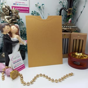 May include: A gold envelope with a flap on the top. The envelope is sitting on a table with a decorative seal and a string of gold beads.