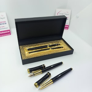 Puede incluir: Un juego de bolígrafos y portaminas negros con detalles dorados, presentado en una caja negra con interior dorado. El juego incluye una pluma estilográfica y un bolígrafo, ambos con adornos dorados.
