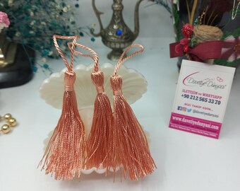 May include: Three peach-colored tassels with a white string attached to each. The tassels are displayed on a white seashell-shaped object.