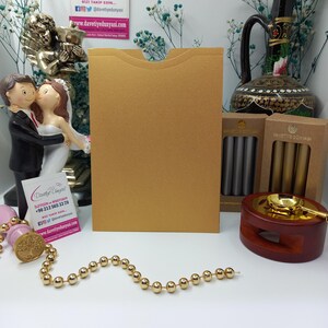May include: A gold envelope with a flap on the top. The envelope is sitting on a white surface with a string of gold beads, a wooden stamp, and two boxes of wax seals. The boxes are labeled "Davetiye D&uuml;nyası" in gold lettering.