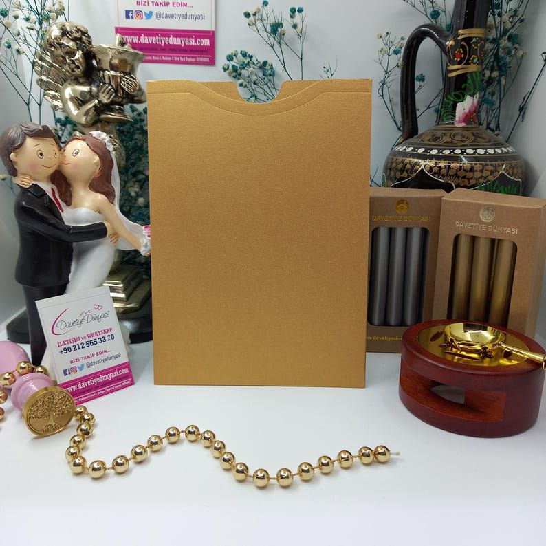 May include: A gold envelope with a flap on the top. The envelope is sitting on a white surface with a string of gold beads, a wooden stamp, and two boxes of wax seals. The boxes are labeled "Davetiye D&uuml;nyası" in gold lettering.