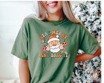 May include: Olive green t-shirt with a retro Santa Claus graphic. The graphic includes the text "Stay Merry and Bright" with a Santa face wearing sunglasses and peace signs. The shirt is a casual, short-sleeved style.