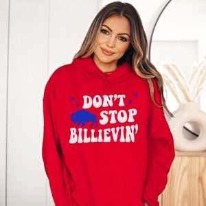 Puede incluir: Sudadera con capucha roja con un gr&aacute;fico blanco que dice "Don't Stop Billievin'" con una silueta de b&uacute;falo azul.