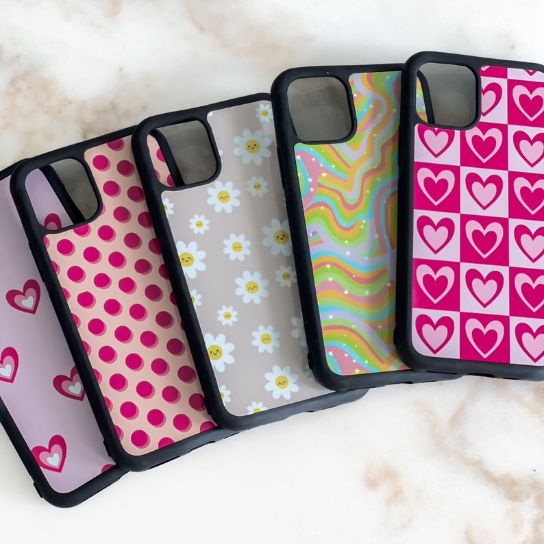 May include: Five different phone cases with various patterns. The cases are black with a clear back and feature pink and white designs. The designs include polka dots, daisies, rainbow swirls, and hearts.