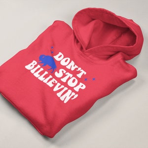 Puede incluir: Sudadera con capucha roja con texto blanco que dice "Don't Stop Believin'" y una silueta de b&uacute;falo azul.