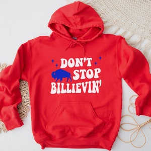 Puede incluir: Sudadera con capucha roja con un gr&aacute;fico blanco que dice "Don't Stop Billievin'" y una silueta azul de un b&uacute;falo.