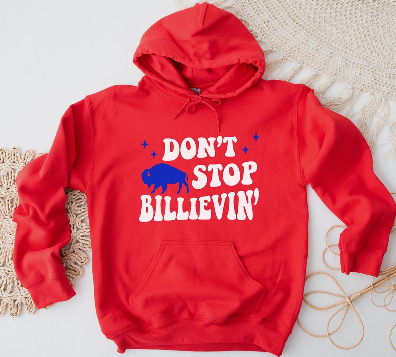 Puede incluir: Sudadera con capucha roja con un gr&aacute;fico blanco que dice "Don't Stop Billievin'" y una silueta azul de un b&uacute;falo.