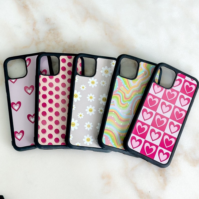 May include: Five different phone cases with various patterns. The cases are black with pink and white designs. The designs include hearts, polka dots, flowers, and wavy lines.