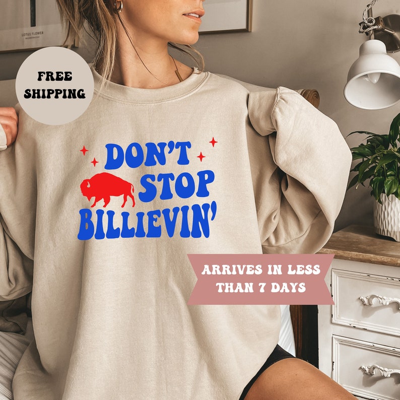 May include: A beige sweatshirt with a red buffalo and blue text that reads "Don't Stop Billievin'" printed on the front.