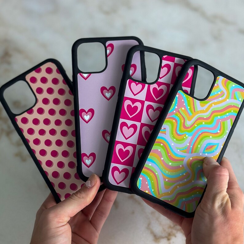 May include: Four different phone cases with various patterns. The cases are black with pink, white, and blue designs. One case has a polka dot pattern, another has a checkered pattern with hearts, another has a swirl pattern, and the last one has a heart pattern.