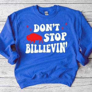 Puede incluir: Una sudadera azul real con texto blanco que dice "Don't Stop Believin'" en una fuente retro. Una silueta roja de un b&uacute;falo est&aacute; por encima del texto.