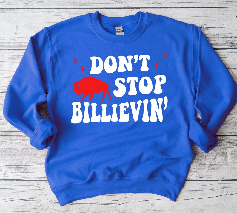 Puede incluir: Una sudadera azul real con texto blanco que dice "Don't Stop Believin'" en una fuente retro. Una silueta roja de un b&uacute;falo est&aacute; por encima del texto.