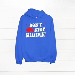 Puede incluir: Una sudadera con capucha azul real con un gr&aacute;fico blanco que dice "Don't Stop Billievin'" con una silueta de b&uacute;falo roja.