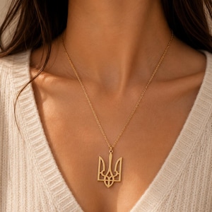 May include: A gold-toned necklace featuring a pendant shaped like the Ukrainian Tryzub, also known as the trident symbol. The pendant is suspended from a delicate gold-toned chain that rests against the collarbone, showcasing a polished finish.