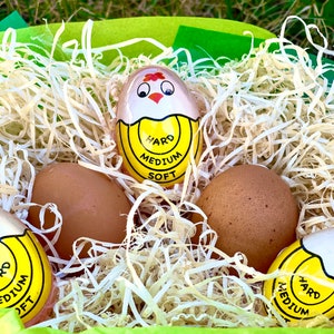 May include: A basket filled with brown eggs and three yellow and white egg-shaped timers with a chicken face and the words "Hard", "Medium", and "Soft" printed on them.