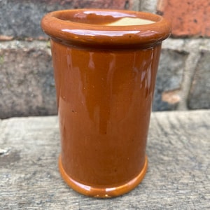 Puede incluir: Un tarro cilíndrico de cerámica marrón con acabado brillante. El tarro tiene un borde y una base redondeados, y un interior ligeramente más claro. El exterior es de color marrón cálido y uniforme. Está sobre una superficie de madera envejecida.