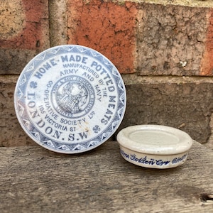 May include: A vintage, white ceramic lid with blue lettering that reads "Home Made Potted Meats, Manufactured by the Army and Navy Co-operative Society Ltd, 105 Victoria St, London, S.W."