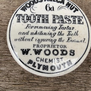 Peut inclure: Un récipient rond blanc cassé avec du texte noir. Le texte indique "WOODS ARECA NUT TOOTH PASTE" et décrit son objectif. Le récipient indique également "W. WOODS CHEMIST PLYMOUTH".