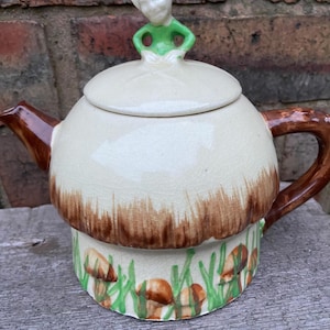 Peut inclure: Une théière en céramique fantaisiste en forme de champignon. Le corps est crème avec un chapeau et une anse marron. Une petite figurine d'une personne en vert et blanc est sur le couvercle. La base est décorée de champignons peints et d'herbe verte.