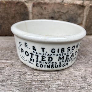 May include: A white ceramic pot with black printed text that reads R. & T. GIBSON MANUFACTURERS OF POTTED MEATS 93 PRINCES STREET EDINBURGH. The small, round container features a thick rim and a smooth, off-white finish with visible aging.