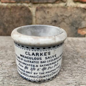 May include: A small, antique ceramic jar with a white base and a black and white patterned border. The jar features the text "CLARKES MIRACULOUS SALVE" and other text. The jar is sitting on a wooden surface.