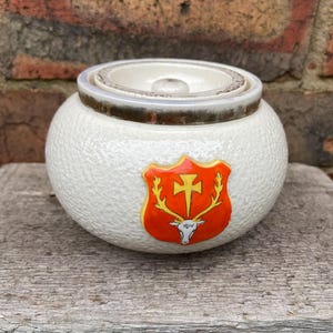Può includere: Vaso in ceramica bianca con coperchio e superficie strutturata. Presenta un emblema decorativo con una croce e corna di cervo in rosso, giallo e bianco. Il vaso è appoggiato su una superficie di legno.