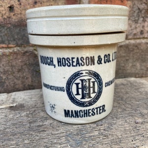 Pode incluir: Um pote de cerâmica branco com uma tampa. O pote tem letras pretas que dizem "Hough, Hoseason & Co. Ltd. Manufacturing Chemists Manchester" e um logotipo preto com as letras "HH" dentro de uma coroa.