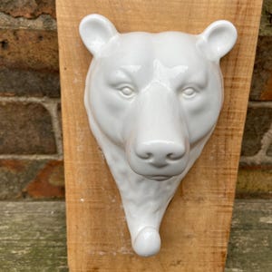 Può includere: Un gancio da parete a forma di testa di orso in ceramica bianca montato su una tavola di legno. La testa dell'orso presenta dettagli e un gancio nella parte inferiore. La tavola di legno ha una venatura naturale. Il gancio è adatto per appendere oggetti.