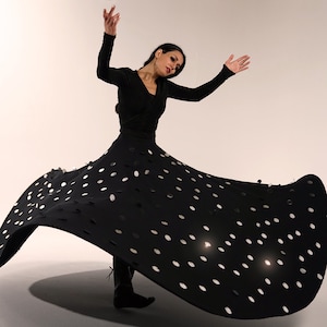 Puede incluir: Una bailarina con un vestido negro con lunares blancos. El vestido de manga larga se ensancha dramáticamente, creando una forma dinámica. Los brazos de la bailarina están levantados y lleva botas negras.