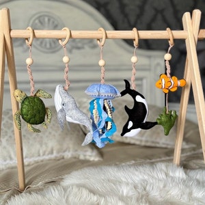 以下が含まれることがあります： 木製のベビー プレイジムに、5つのフェルトのおもちゃがぶら下がっています。緑のウミガメ、青いクジラ、青いクラゲ、白黒のシャチ、オレンジと白のクマノミです。