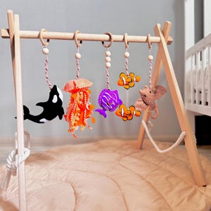 Puede incluir: Gimnasio para bebés de madera con juguetes colgantes con temática oceánica. Los juguetes incluyen una orca negra y blanca, una medusa naranja, un pez morado, dos peces payaso naranjas y un elefante marrón. El gimnasio está sobre una alfombra rosa.