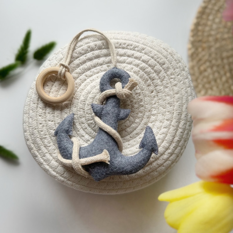 May include: A gray felt anchor toy with a cream-colored rope is displayed on a round, woven basket. A wooden ring is attached to the top of the anchor with a cream-colored rope. The anchor is a nautical-themed item.
